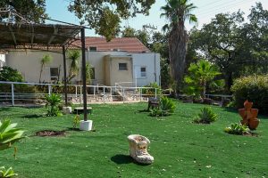 בית לבן עם גג אדום וגינה ירוקה גדולה עם צמחיה וקישוטים.
